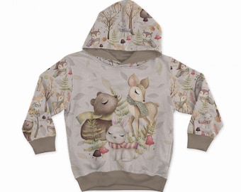 Peut inclure: Un sweat à capuche beige clair avec un imprimé intégral d'animaux des bois, dont un ours, un cerf et un lapin portant des écharpes, entourés d'arbres, de fougères et de champignons. Le vêtement a des manches longues avec des poignets marron.