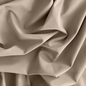 Könnte beinhalten: Nahaufnahme eines beigefarbenen Stoffes mit weichen Falten und subtilen Schatten. Der Stoff scheint ein glattes, gewebtes Material zu sein, möglicherweise Baumwolle oder eine ähnliche Mischung. Die neutrale Farbe und Textur deuten auf eine Verwendung für Kleidung oder Heimdekoration hin.