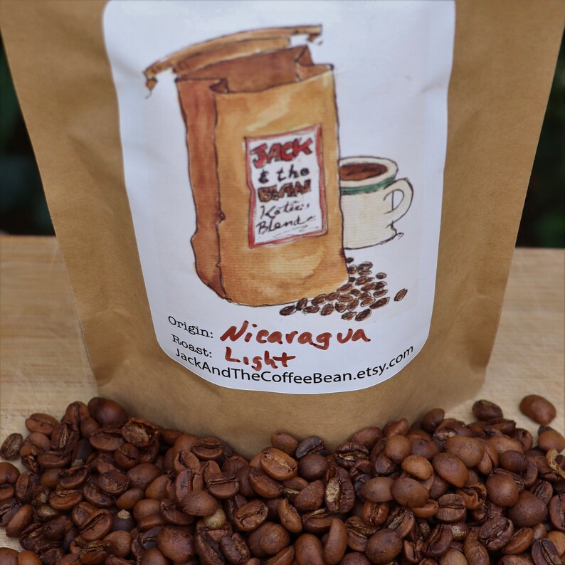 May include: A brown paper bag with a white label. The label has a hand-drawn illustration of a coffee bag with the text "Jack & the Bean Katie's Blend" and a cup of coffee. The label also includes the text "Origin: Nicaragua Roast: Light JackAndTheCoffeeBean.etsy.com". The bag is sitting on a pile of coffee beans.