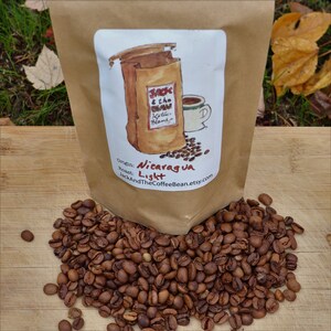 May include: A brown paper bag with a white label that says "Jack & the Bean, Kaitlin Blend, Origin: Nicaragua, Roast: Light, JackAndTheCoffeeBean.etsy.com". The bag is open and a pile of roasted coffee beans are spilling out.