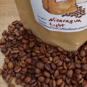 May include: A brown paper bag with a white label that says "Origin: Nicaragua Roast: Light JackAndTheCoffeeBean.etsy.c" and a pile of brown coffee beans spilling out of the bag.