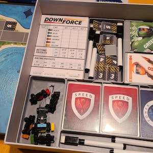 May include: A game board featuring a racetrack and a box of game pieces. The box includes a selection of game pieces, such as cars, dice, and cards. The game is called "Downforce".