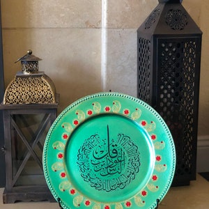 May include: A teal charger plate with a gold rim and red and gold accents. The plate features a black Arabic calligraphy design in the center.