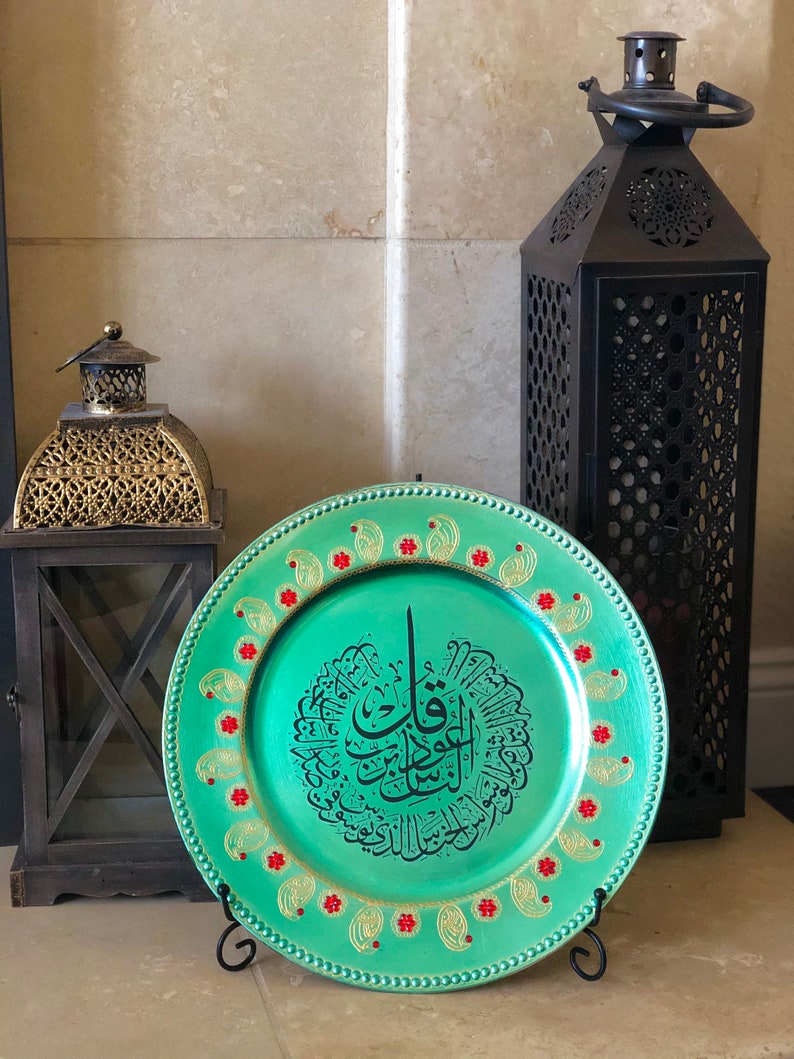 May include: A teal charger plate with a gold rim and red and gold accents. The plate features a black Arabic calligraphy design in the center.