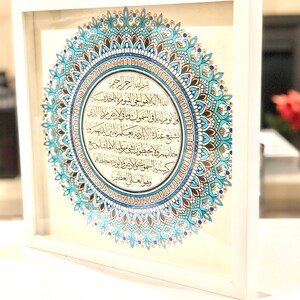 May include: A framed print of a mandala with Arabic calligraphy in a white frame. The mandala is blue, gold, and red with intricate details. The calligraphy is in black ink and reads "الد اله الا هو الحي القيوم ا نا خلا سنة لو نوم له يا في السموات وما في ارض مرزا الذي يشفع عنده البادية يعلم ما من الدهر و شهر ولا يحيطون بو مرعليه الاماشاء 350 كسية السموت والارض و الورا احفظهم وهو العلى العظ".