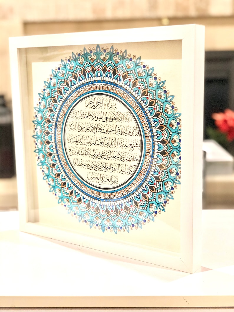 May include: A framed print of a mandala with Arabic calligraphy in a white frame. The mandala is blue, gold, and red with intricate details. The calligraphy is in black ink and reads "الد اله الا هو الحي القيوم ا نا خلا سنة لو نوم له يا في السموات وما في ارض مرزا الذي يشفع عنده البادية يعلم ما من الدهر و شهر ولا يحيطون بو مرعليه الاماشاء 350 كسية السموت والارض و الورا احفظهم وهو العلى العظ".