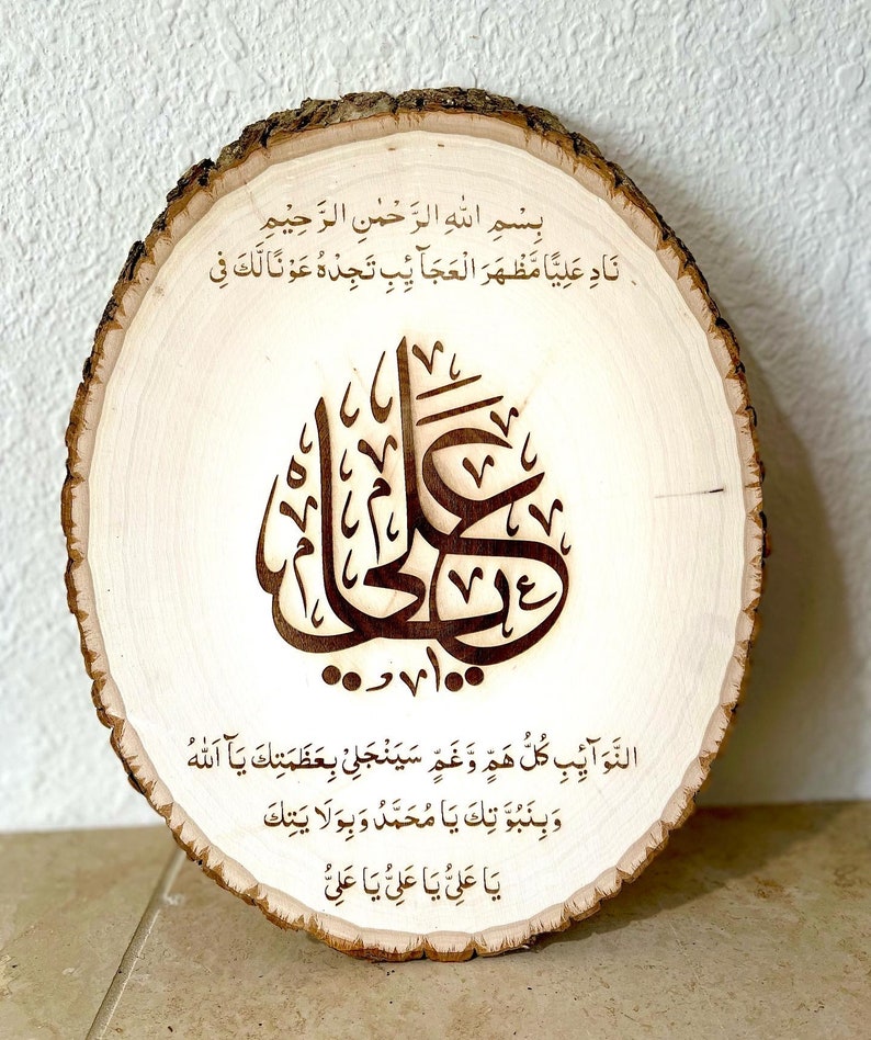 May include: A wooden round slice with a brown wood grain pattern. The slice has an inscription in Arabic script, which reads "Bismillah ir-Rahman ir-Rahim Nad Aliya Mazhara al-Ajaib Tajede Awna Laka Fi al-Nawaib Kullu Haza Wa Gham Sayanjalli Bi-Azamatik Ya Allah Wa Binubuwwatik Ya Muhammad Wa Bi-Wilayatik Ya Ali Ya Ali Ya Ali".