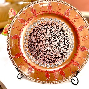 May include: A decorative round plate with a gold background and red and white accents. The center of the plate features a circular design with black Arabic calligraphy.