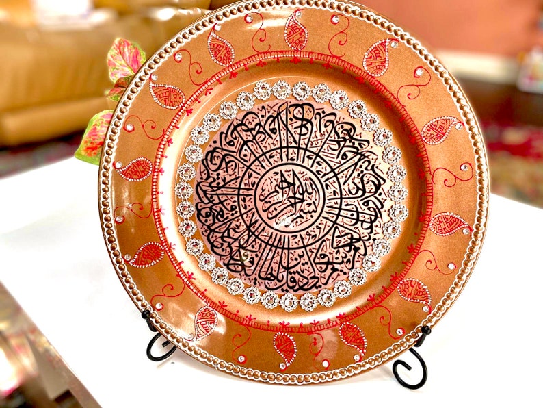 May include: A decorative round plate with a gold background and red and white accents. The center of the plate features a circular design with black Arabic calligraphy.