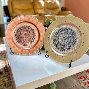 May include: Two decorative charger plates with intricate black calligraphy designs on a white surface. One plate is gold with black calligraphy and the other is peach with black calligraphy.
