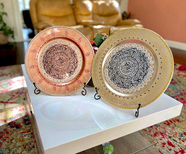 May include: Two decorative charger plates with intricate black calligraphy designs on a white surface. One plate is gold with black calligraphy and the other is peach with black calligraphy.