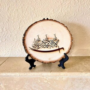 May include: A wooden round sign with a black Arabic calligraphy design on a natural wood background. The sign is on a black stand.