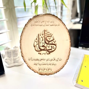May include: A wooden round plaque with a decorative stand. The plaque features a carved Arabic inscription in brown, with the text "Bismillah ir-Rahman ir-Rahim Nad Aliya Mazhar al-Ajaib Tijdu Awna Laka Fi..."  The inscription is surrounded by a decorative border.