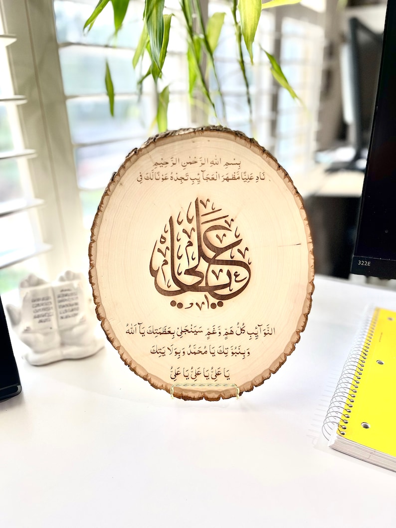 May include: A wooden round plaque with a decorative stand. The plaque features a carved Arabic inscription in brown, with the text "Bismillah ir-Rahman ir-Rahim Nad Aliya Mazhar al-Ajaib Tijdu Awna Laka Fi..."  The inscription is surrounded by a decorative border.