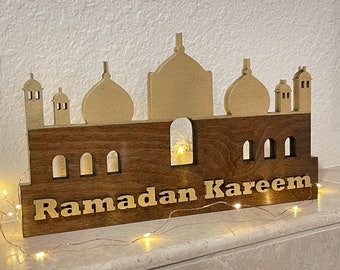 May include: A wooden Ramadan Kareem decoration featuring a mosque silhouette with gold-colored domes and minarets. The base is a dark brown color with the words "Ramadan Kareem" cut out. A string of warm white lights is wrapped around the base.
