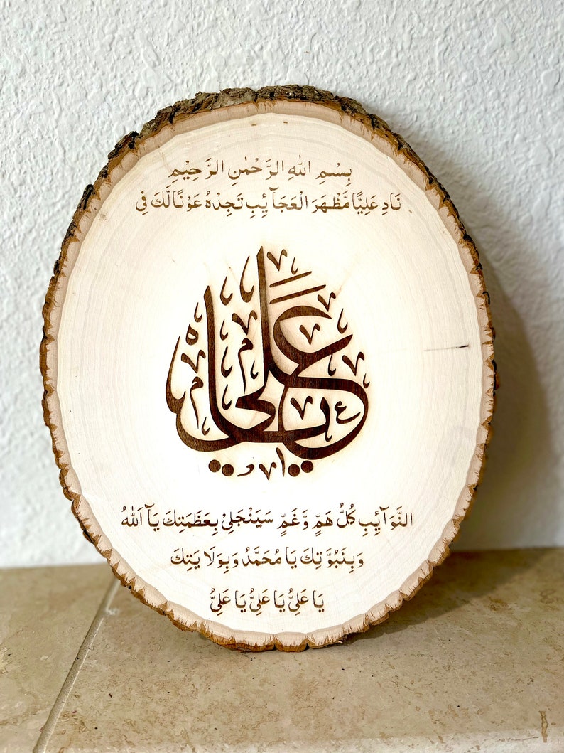 May include: A wooden round slice with a burned-in Arabic calligraphy design. The text reads "Bismillah al-Rahman al-Rahim Nad'a Aliya Mazhara al-'aja'ib Tajidhu 'awnan Laka Fi al-nawa'ib Kullu Haa Wa Gha'a Sayanjal'i Bi'azamat'ika Ya Allah Wa binubuwwat'ika Ya Muhammad Wa biwilayat'ika Ya Ali Ya Ali Ya Ali"