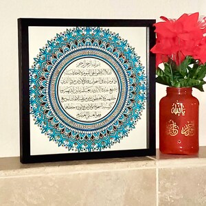 May include: A framed black and white print with a blue and silver mandala design. The print features Arabic calligraphy in a circle. The print is on a shelf with a red vase with gold Arabic calligraphy.