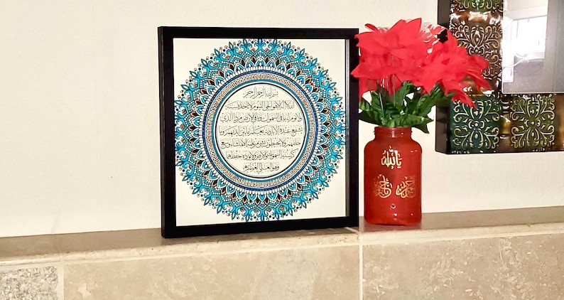 May include: A framed black and white print with a blue and silver mandala design. The print features Arabic calligraphy in a circle. The print is on a shelf with a red vase with gold Arabic calligraphy.