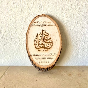 May include: A wooden round slice with Arabic calligraphy in brown. The text reads "Bismillah al-Rahman al-Rahim Nadin Aliyan Mazhara al-Ajaib Tjidhu Awna Laka Fi 100 Dawah Al-Nawaib Kullu Hamm Wa Gham Sayanjalli Bi Azamatka Ya Allah Wa Binubuwatika Ya Muhammad Wa Bi Wilattika Ya Ali Ya Ali Ya Ali".