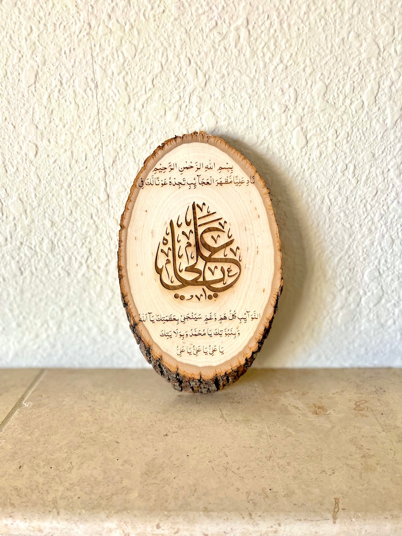 May include: A wooden round slice with Arabic calligraphy in brown. The text reads "Bismillah al-Rahman al-Rahim Nadin Aliyan Mazhara al-Ajaib Tjidhu Awna Laka Fi 100 Dawah Al-Nawaib Kullu Hamm Wa Gham Sayanjalli Bi Azamatka Ya Allah Wa Binubuwatika Ya Muhammad Wa Bi Wilattika Ya Ali Ya Ali Ya Ali".