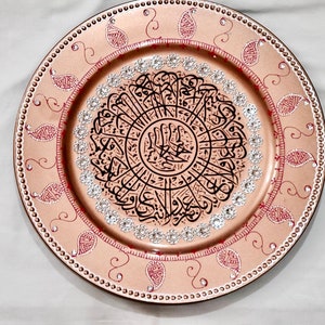 May include: A decorative round plate with a gold background and red and white paisley designs. The center of the plate features a circular design with black Arabic calligraphy and silver rhinestones.