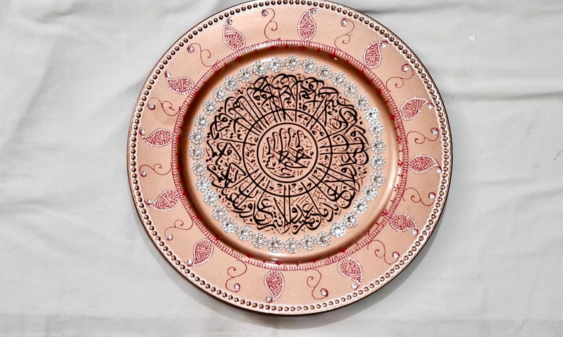 May include: A decorative round plate with a gold background and red and white paisley designs. The center of the plate features a circular design with black Arabic calligraphy and silver rhinestones.