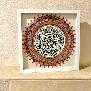 May include: A framed artwork featuring a circular mandala design with intricate details in shades of orange, red, and purple. The mandala is surrounded by a white frame and features Arabic calligraphy in black.
