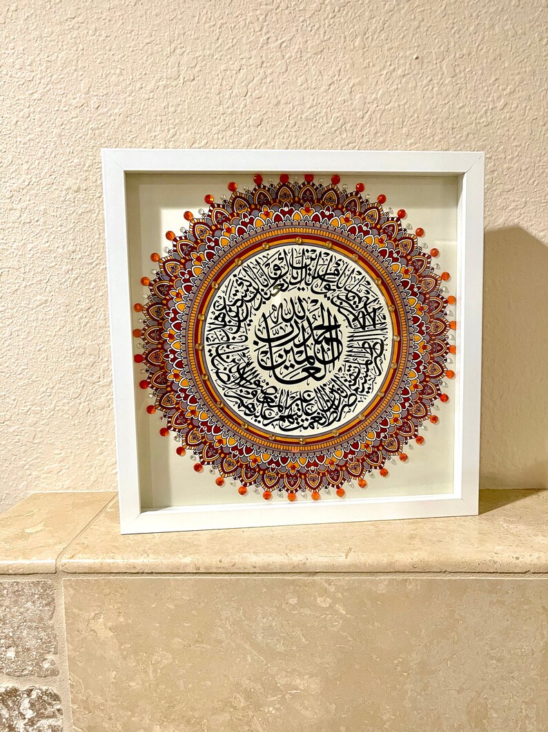 May include: A framed artwork featuring a circular mandala design with intricate details in shades of orange, red, and purple. The mandala is surrounded by a white frame and features Arabic calligraphy in black.