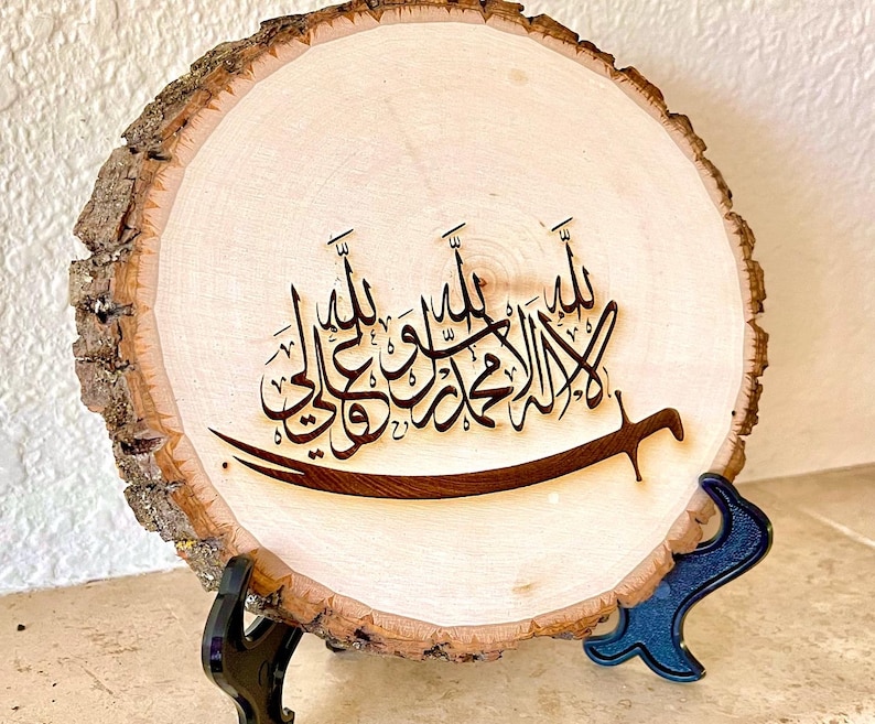 May include: A wooden round slice with a black stand, featuring a wood-burned Arabic calligraphy design. The calligraphy reads "Allah", "Muhammad", and "Ali" in Arabic.