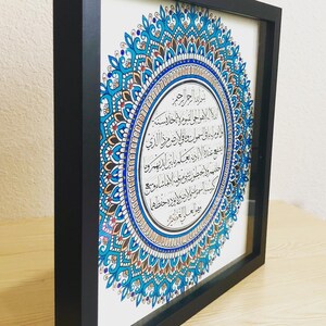 May include: A framed artwork featuring a blue and gold mandala with intricate details. The mandala surrounds a white circle with Arabic calligraphy.