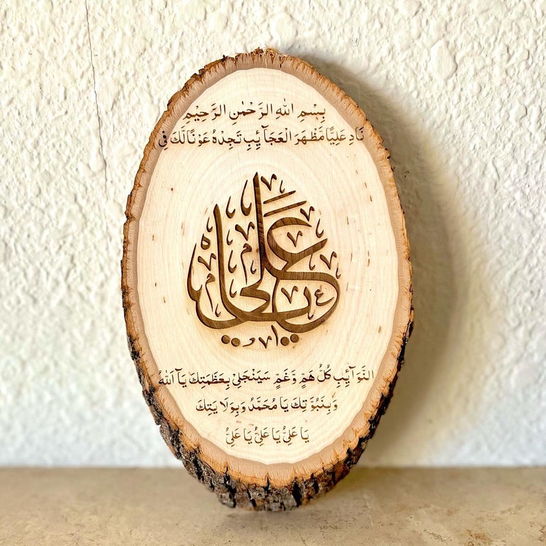 May include: A wooden round plaque with Arabic calligraphy and text in brown. The text reads "Bismillah ir-Rahman ir-Rahim Nad Aliya Mazhar al-Ajaib Tajedeh Awna Laka Fi 100 Da'wa Al-Nawaib Kullu Ham Wa Ghamah Sayanjalli Bi'azamataka Ya Allah Wa Binubuwwatoka Ya Muhammad Wa Biwilayatika Ya Ali Ya Ali Ya Ali."