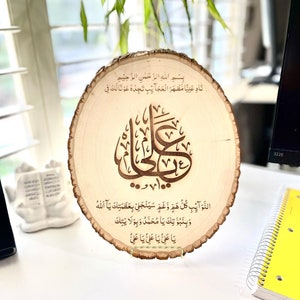 May include: A wooden plaque with a circular shape and a rough-hewn edge. The plaque features a decorative design of Arabic calligraphy in brown, with the text "Bismillah ir-Rahman ir-Rahim Nad Aliya Mazhar al-Ajaib Tajidhu Awna Laka Fi Al-Nawaib Kullu Huwa Wa Gam Sayanjalli Bi-Azamatik Ya Allah Wa Binabuwatik Ya Muhammad Wa Bi-Wilayatik Ya Ali Ya Ali Ya Ali" inscribed on the plaque.