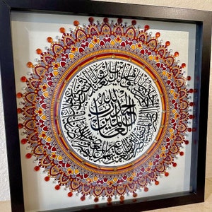May include: A framed mandala with intricate details in shades of red, orange, and gray. The center of the mandala features a circular design with Arabic calligraphy in black ink.
