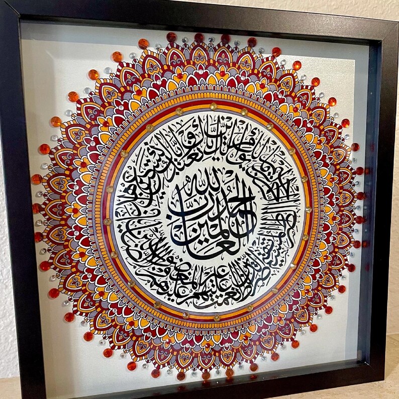 May include: A framed mandala with intricate details in shades of red, orange, and gray. The center of the mandala features a circular design with Arabic calligraphy in black ink.