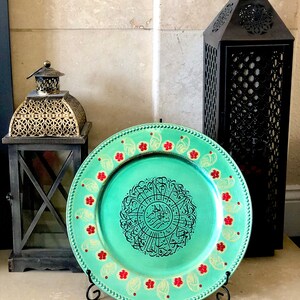 May include: A decorative charger plate with a teal green background and gold accents. The center of the plate features a circular design with black Arabic calligraphy. The plate is surrounded by a border of red and gold flowers.