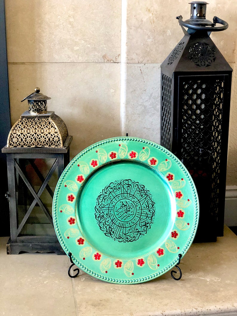 May include: A decorative charger plate with a teal green background and gold accents. The center of the plate features a circular design with black Arabic calligraphy. The plate is surrounded by a border of red and gold flowers.