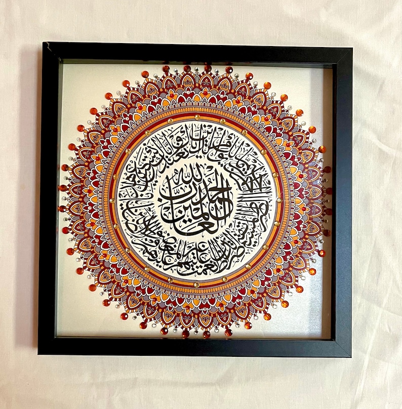 May include: A framed artwork featuring a circular mandala design with intricate details in shades of red, gold, and brown. The center of the mandala contains a calligraphic inscription in black.