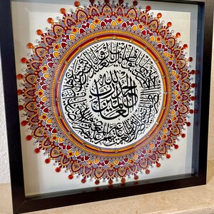 May include: A framed artwork featuring a mandala design with intricate details in shades of red, orange, and gray. The center of the mandala features a circular inscription in Arabic calligraphy.