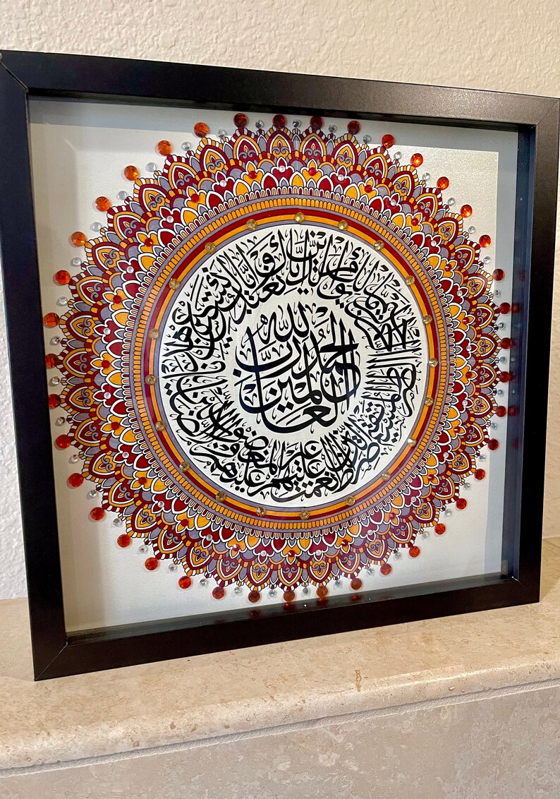 May include: A framed artwork featuring a mandala design with intricate details in shades of red, orange, and gray. The center of the mandala features a circular inscription in Arabic calligraphy.