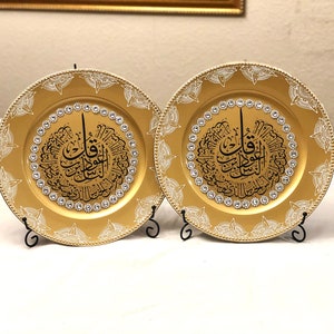 May include: Two gold charger plates with a white and black design featuring Arabic calligraphy and rhinestones. The plates are on black metal stands.