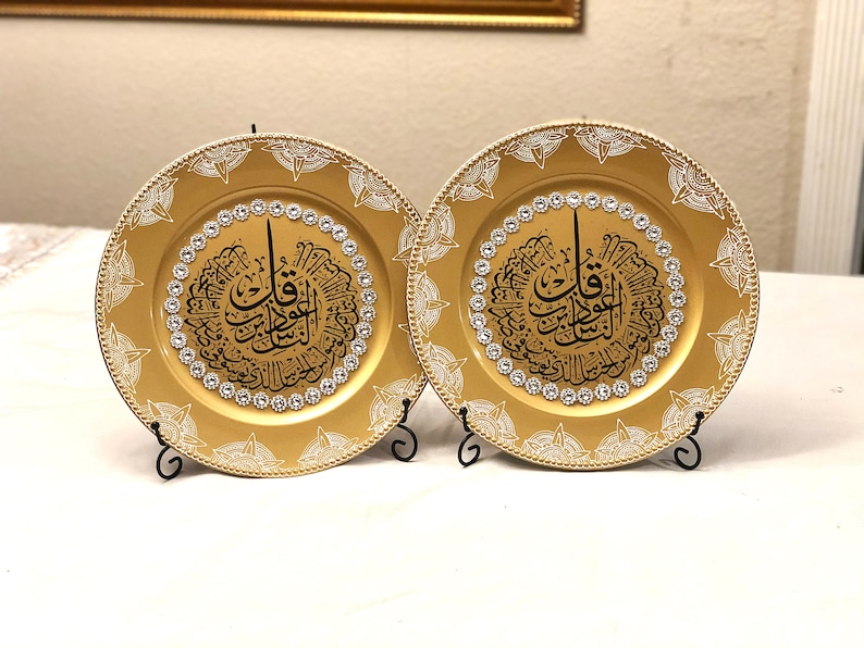 May include: Two gold charger plates with a white and black design featuring Arabic calligraphy and rhinestones. The plates are on black metal stands.