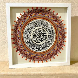 May include: A framed artwork featuring a colorful mandala design with intricate details. The mandala is surrounded by a white border and is set against a white background. The center of the mandala features Arabic calligraphy in black.