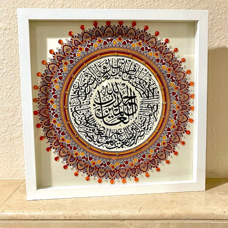 May include: A framed artwork featuring a colorful mandala design with intricate details. The mandala is surrounded by a white border and is set against a white background. The center of the mandala features Arabic calligraphy in black.