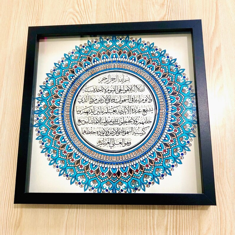 May include: A framed artwork featuring a circular mandala design in shades of blue, gold, and red. The center of the mandala contains an inscription in Arabic script.