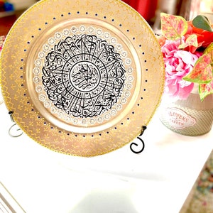 May include: A decorative round plate with a gold rim and blue accents. The center of the plate features a black and white calligraphic design with a silver background. The plate is on a stand.