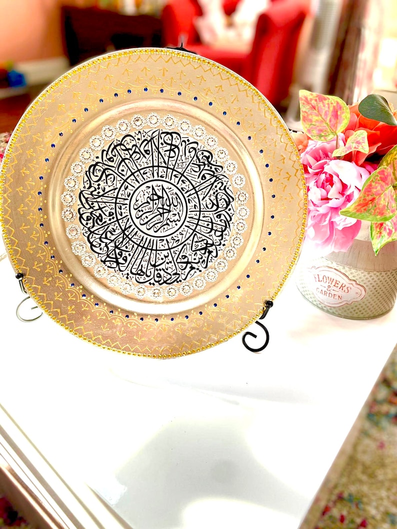 May include: A decorative round plate with a gold rim and blue accents. The center of the plate features a black and white calligraphic design with a silver background. The plate is on a stand.