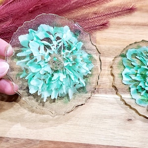 Könnte beinhalten: Zwei runde, transparente Harz-Untersetzer mit einem gewellten Rand. Jeder Untersetzer zeigt eine gepresste, getrocknete, hellgrüne Blume mit goldenen Glitzerakzenten.