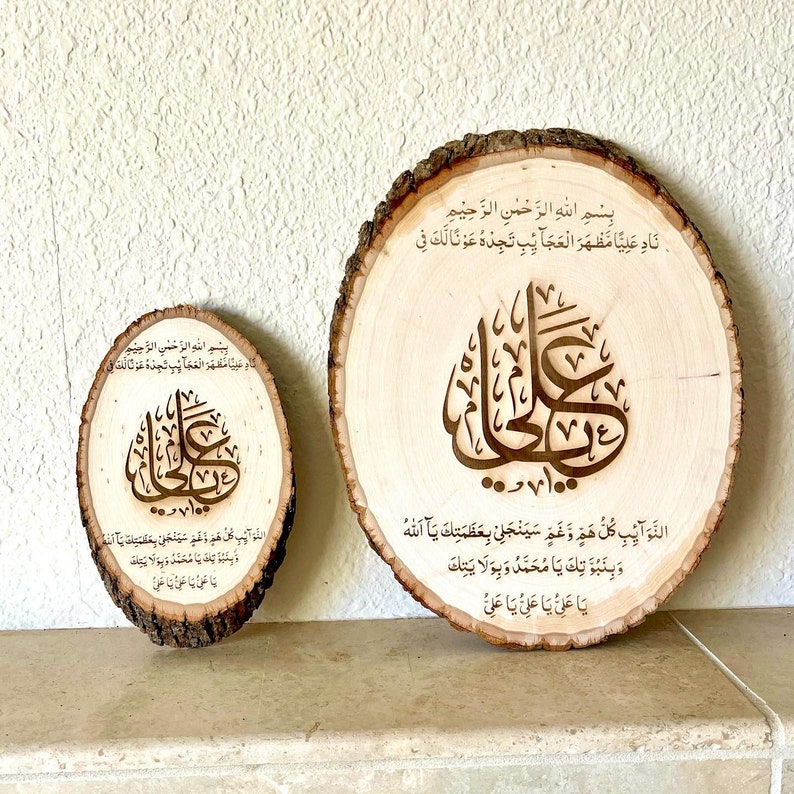 May include: Two wooden slices with Arabic calligraphy and text in a brown wood frame. The larger slice has the text "Bismillah ir-Rahman ir-Rahim Nad Aliya Mazhar al-Ajaib Tijidhu Awna Laka Fi al-Nawaib Kullu Hamm wa Gham Sayanjalli Bi-Azamatka Ya Allah wa Binubuwwatika Ya Muhammad wa Bi-Wilayatika Ya Ali Ya Ali Ya Ali 100 Decade" The smaller slice has the text "Bismillah ir-Rahman ir-Rahim Ya Ali Ya Ali Ya Ali"