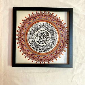 May include: A framed artwork featuring a circular mandala design with intricate details in shades of red, orange, and gold. The center of the mandala contains Arabic calligraphy in black ink.