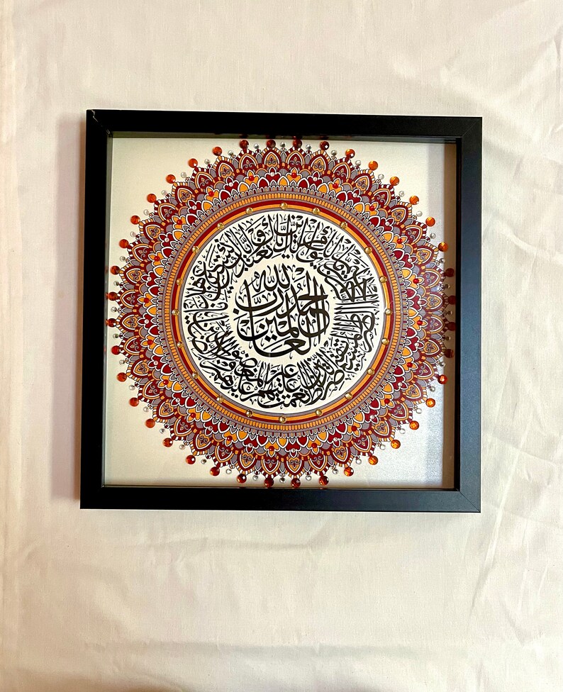 May include: A framed artwork featuring a circular mandala design with intricate details in shades of red, orange, and gold. The center of the mandala contains Arabic calligraphy in black ink.